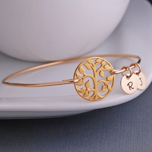 Puede incluir: Pulsera de brazalete dorada con un dije de árbol de la vida y dos dijes con iniciales. Las iniciales son R y J.