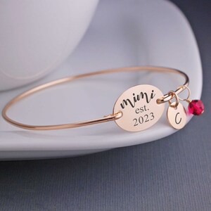May include: A rose gold bangle bracelet with a charm that reads "Mimi est. 2023". A small charm with the letter "C" and a red crystal dangle from the bracelet.