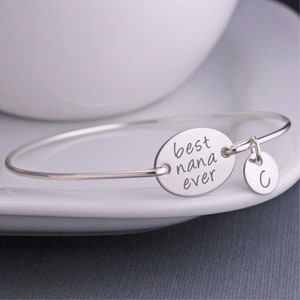 May include: Silver bangle bracelet with a charm that says "best nana ever" and a letter "C" charm.