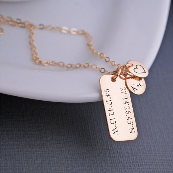 May include: A gold necklace with a rectangular charm engraved with the coordinates 27°14'26.45"N 94°17'42.15"W. The necklace also has two smaller charms, one with the initials "K+C" and the other a heart.