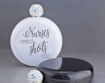 May include: Two round, silver-colored hip flasks with a crystal-like stopper. One flask is white with the text "Nurses need shots too". The other flask is black with a sparkly finish.
