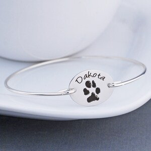 May include: Silver bangle bracelet with a silver oval charm engraved with the name "Dakota" and a paw print.