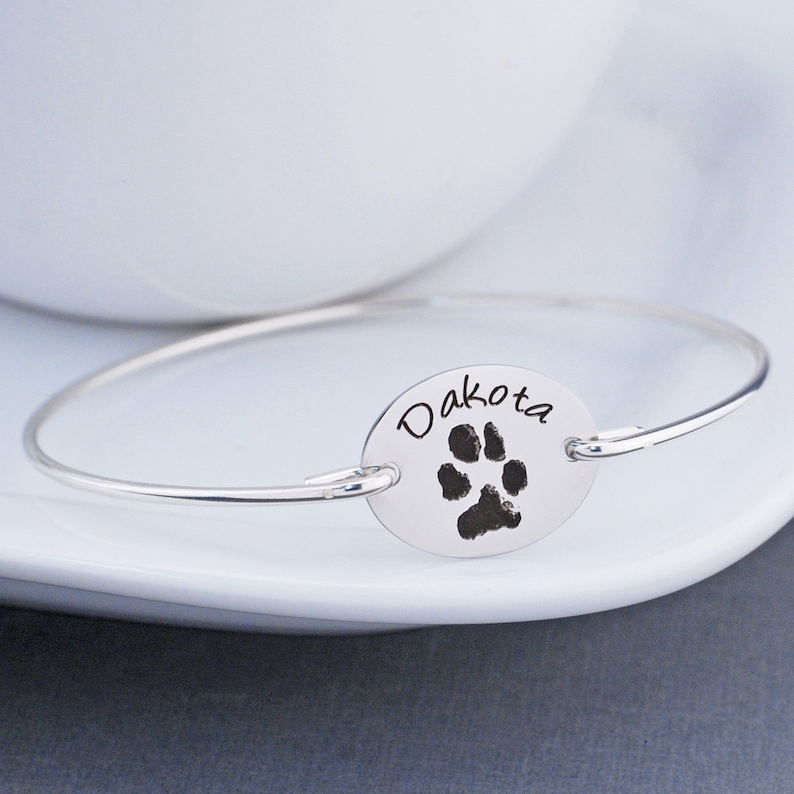 May include: Silver bangle bracelet with a silver oval charm engraved with the name "Dakota" and a paw print.