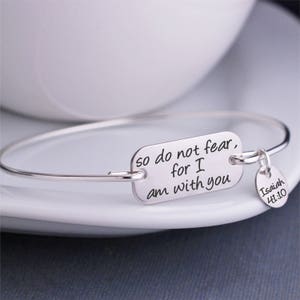 May include: Silver bracelet with a rectangular charm that reads "so do not fear, for I am with you". A smaller round charm with the text "Isaiah 41:10" hangs from the bracelet.