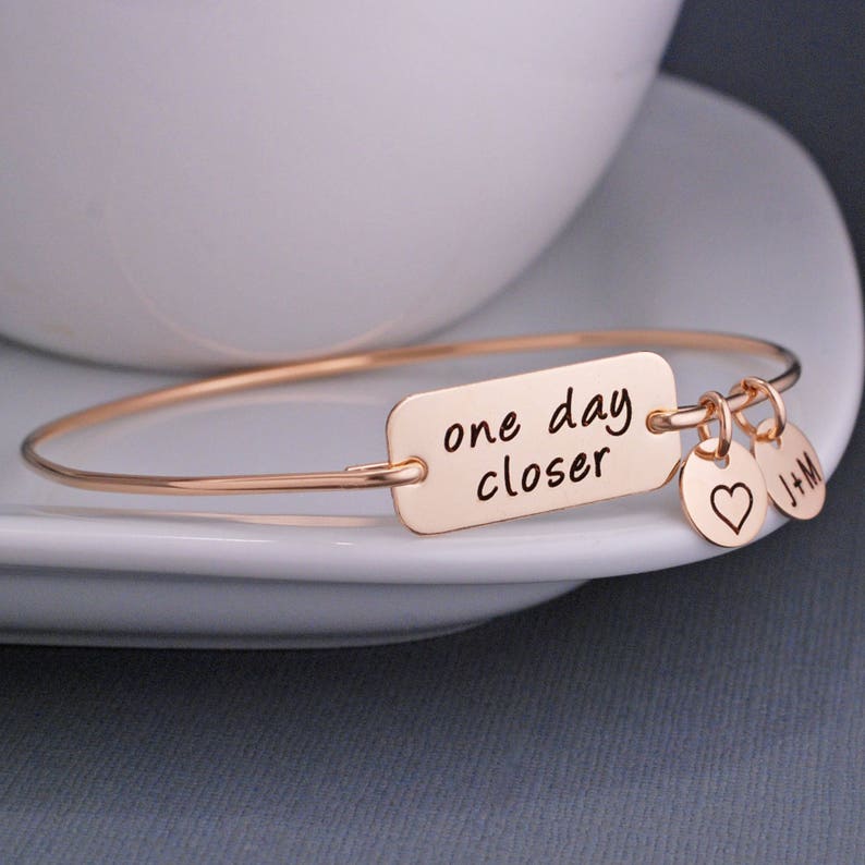 May include: Rose gold expandable bracelet with a rectangular charm that reads "one day closer". The bracelet has two small charms attached, one with a heart and the other with the initials "J+M".