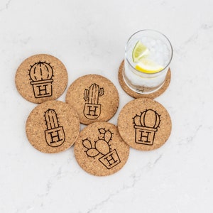 Può includere: Sei sottobicchieri in sughero con un design a cactus. Ogni sottobicchiere presenta una silhouette di cactus diversa con la lettera "H" al centro. I sottobicchieri sono disposti in cerchio attorno a un bicchiere d'acqua con ghiaccio e una fetta di limone.