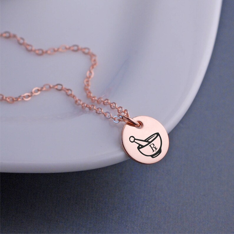 May include: A rose gold necklace with a round pendant featuring a mortar and pestle with the letter "R" inside.
