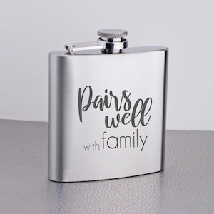 Może przedstawiać: Srebrna, stalowa flaszka z zawiasem. Na przodzie flaszki wygrawerowano napis "Pairs well with family".