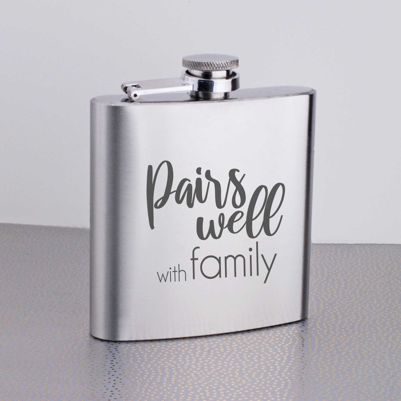 Może przedstawiać: Srebrna, stalowa flaszka z zawiasem. Na przodzie flaszki wygrawerowano napis "Pairs well with family".
