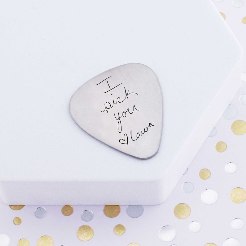 Handwritten message engraved on a guitar pick.  Message says I pick you heart symbol laura
made of stainless steel and measures 1.25 inches by 1 inch