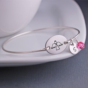 K&ouml;nnte beinhalten: Silbernes Armreif-Armband mit einem ovalen Anh&auml;nger mit einer Engelskontur und einem kleinen Anh&auml;nger mit dem Buchstaben 'S' und einem rosa Kristall.