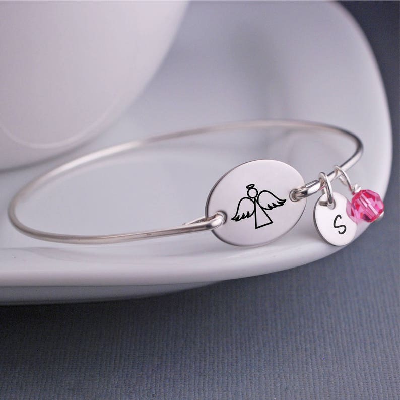 K&ouml;nnte beinhalten: Silbernes Armreif-Armband mit einem ovalen Anh&auml;nger mit einer Engelskontur und einem kleinen Anh&auml;nger mit dem Buchstaben 'S' und einem rosa Kristall.
