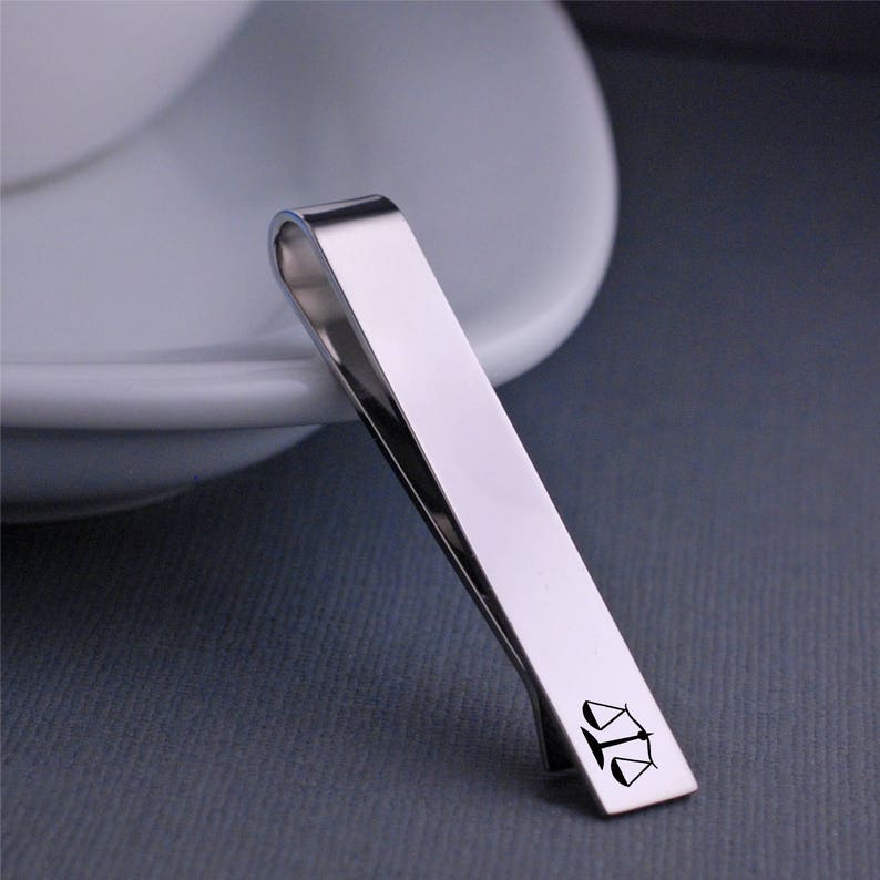 Puede incluir: Pinza de corbata plateada con un dise&ntilde;o de balanza de la justicia.