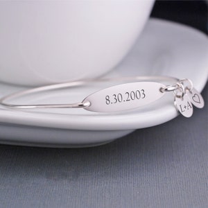 May include: Silver bangle bracelet with a charm that says "L+A" and a heart charm. The bracelet also has an oval tag engraved with the date "8.30.2003".