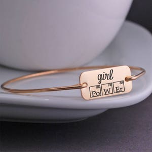 May include: A rose gold bangle bracelet with a rectangular charm that says "girl" and features the chemical symbols for Polonium, Tungsten, and Erbium.