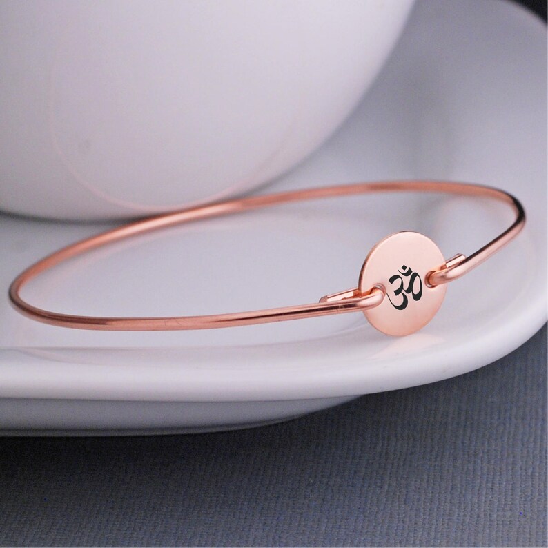 May include: A rose gold bangle bracelet with a small round charm featuring the Om symbol in black.