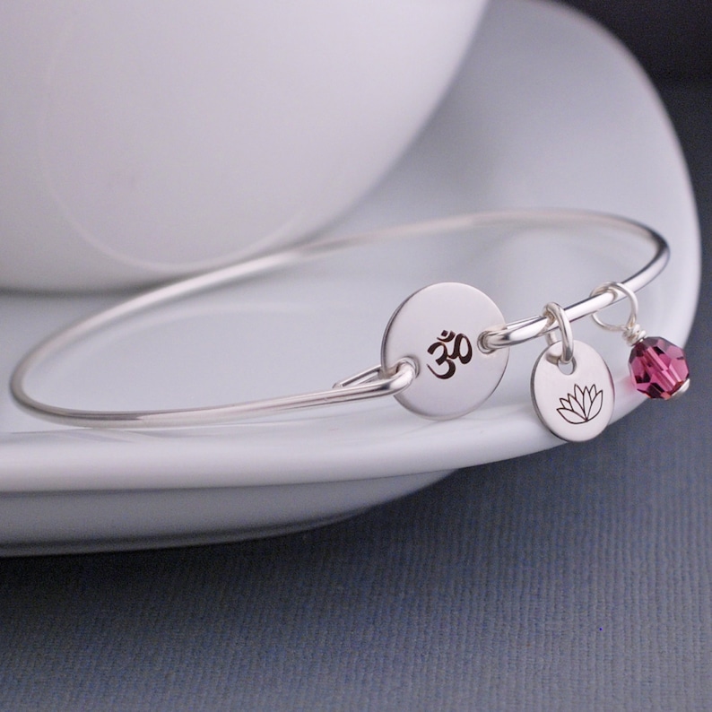 May include: A silver bangle bracelet with a charm featuring the Om symbol and a lotus flower. A small, dark purple crystal dangles from the bracelet.