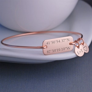 May include: Rose gold bangle bracelet with a rectangular charm engraved with the coordinates 41°56'54.37"N 87°39'19.19"W. The bracelet also has two small charms, one with the letters "L+T" and the other with a heart.
