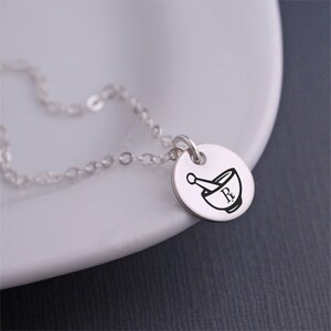 May include: A silver necklace with a round pendant featuring a mortar and pestle with the letter "R" inside. The pendant is engraved with a pharmacy symbol.