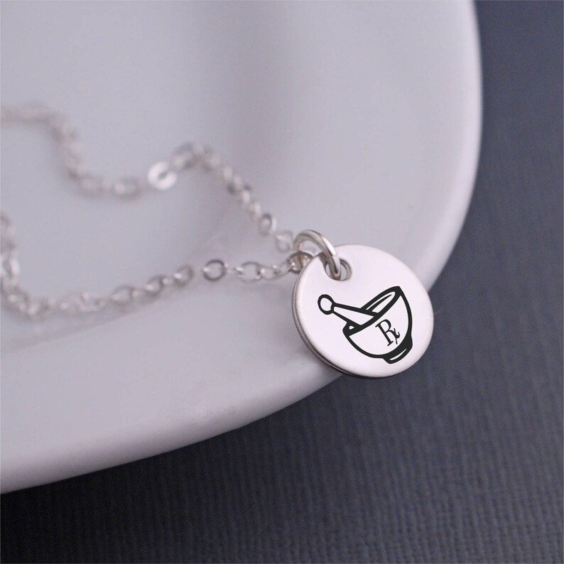 May include: A silver necklace with a round pendant featuring a mortar and pestle with the letter "R" inside. The pendant is engraved with a pharmacy symbol.