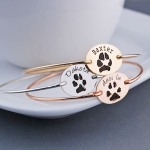 May include: Three silver, gold, and rose gold bracelets with oval charms engraved with paw prints and the names "Dakota", "Baxter", and "Desilu".