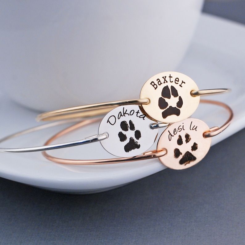 May include: Three silver, gold, and rose gold bracelets with oval charms engraved with paw prints and the names "Dakota", "Baxter", and "Desilu".