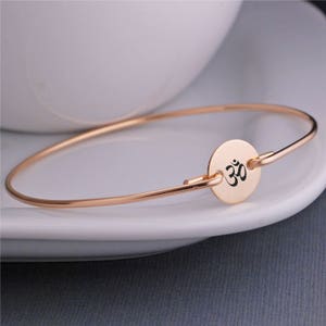 May include: A rose gold bangle bracelet with a round charm featuring the Om symbol in black.