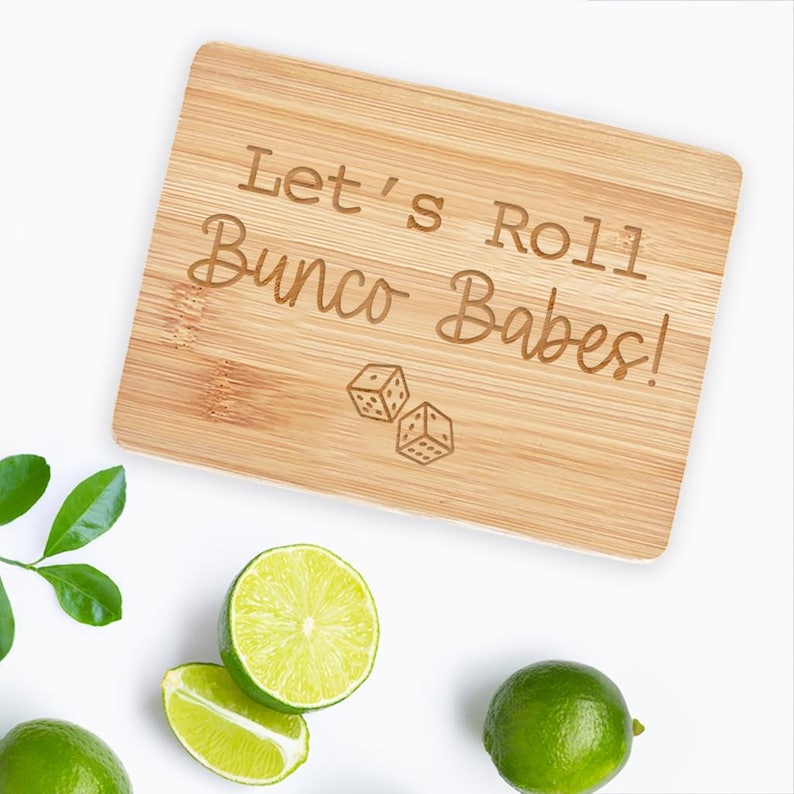 May include: A wooden cutting board with the text "Let's Roll Bunco Babes!" and a pair of dice engraved on it.