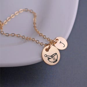 May include: Gold necklace with two charms. One charm is a mortar and pestle with an 'R' inside. The other charm is a letter 'J'.