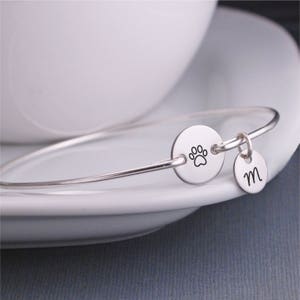 May include: Silver bangle bracelet with a charm featuring a paw print and a charm with the letter M.