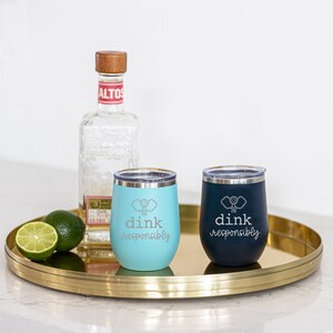 May include: Two stainless steel wine tumblers with a paddle design and the text "dink responsibly". One tumbler is blue and the other is black. The tumblers are on a gold tray with a bottle of tequila in the background.