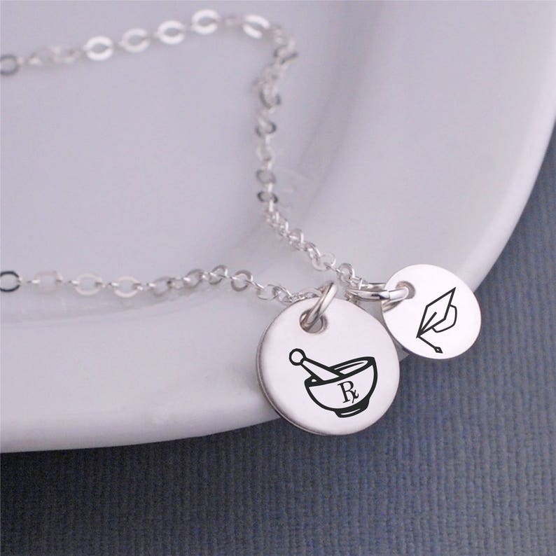 May include: Silver necklace with two charms. One charm features a mortar and pestle with an Rx symbol. The other charm features a graduation cap.
