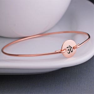 Puede incluir: Un brazalete de oro rosa con un dije redondo que presenta el símbolo Om en negro.