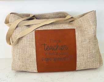 May include: A beige canvas tote bag with brown leather accents. The leather patch has the text "I'm a teacher. What's your superpower?"