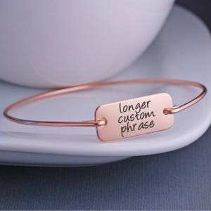 May include: A rose gold bangle bracelet with a rectangular charm that says "longer custom phrase".
