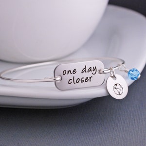 May include: Silver bangle bracelet with a rectangular charm that reads "one day closer" and a small charm with a globe design. The bracelet also has a light blue crystal bead.