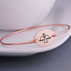 K&ouml;nnte beinhalten: Ein ros&eacute;goldenes Armkettchen mit einem ovalen Anh&auml;nger, der eine einfache Linienzeichnung eines Engels mit Fl&uuml;geln zeigt.