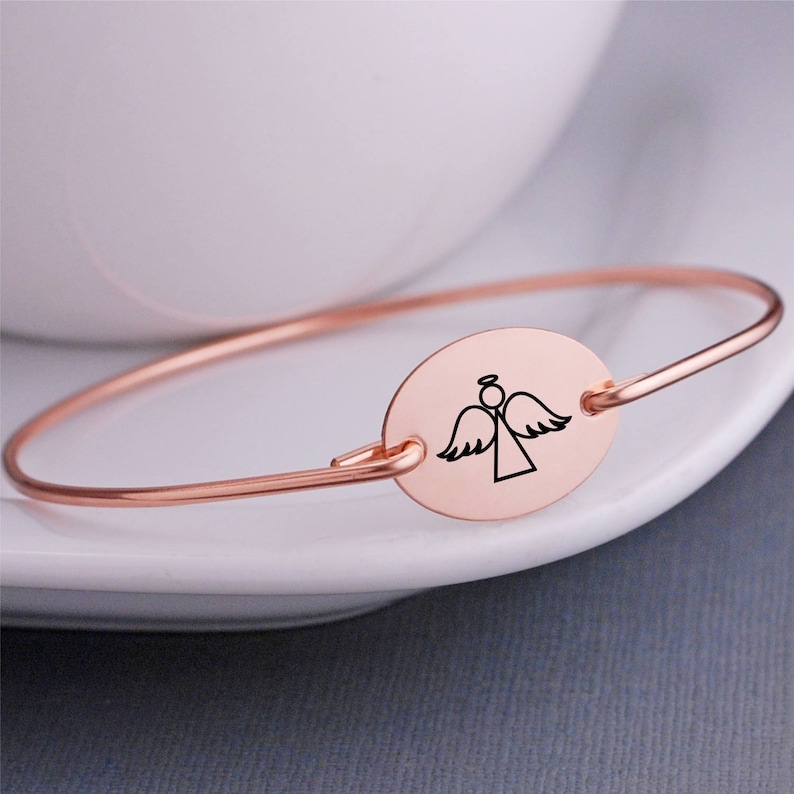 K&ouml;nnte beinhalten: Ein ros&eacute;goldenes Armkettchen mit einem ovalen Anh&auml;nger, der eine einfache Linienzeichnung eines Engels mit Fl&uuml;geln zeigt.