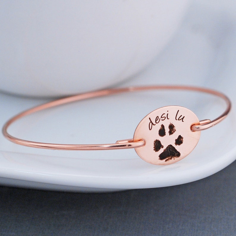 May include: Rose gold bangle bracelet with a personalized oval charm featuring a paw print and the inscription "desi lu".