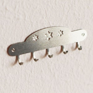 Peut inclure: Un porte-clés en métal argenté avec cinq crochets et un motif décoratif d'étoiles. Le porte-clés est monté sur un mur blanc.