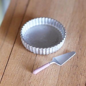 May include: A miniature, silver-colored tart pan with a fluted edge sits on a wooden table. A small, silver pie server with a pink handle is next to the pan. The table has a natural wood grain.