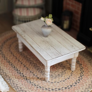May include: A miniature white wooden coffee table with a distressed finish, holding a small grey vase with pink flowers. The table sits on a braided oval rug with neutral colours. The scene evokes a cosy, dollhouse aesthetic.