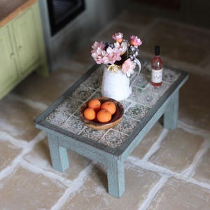 May include: A miniature coffee table with a tiled surface, holding a white jug of flowers, a small bottle of wine, and a wooden bowl of oranges. The table is painted in a distressed blue-green colour.