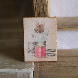 May include: A small wooden sign featuring a vintage illustration of a mouse holding a spool of thread and scissors. The mouse is wearing a red and white striped shirt and has a brown tail.