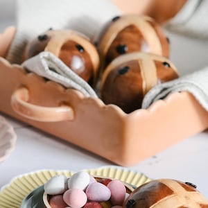 May include: A wooden bowl filled with colourful chocolate Easter eggs sits on a yellow plate with a light blue bowl underneath. A wooden hot cross bun sits next to the bowl.