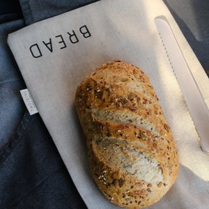 Könnte beinhalten: Ein Sauerteigbrot mit Samen darauf, das auf einem beigefarbenen Leinen-Brotsack mit dem aufgedruckten Wort "BREAD" liegt. Der Sack ist umgeklappt und hat einen grauen Kordelzug.