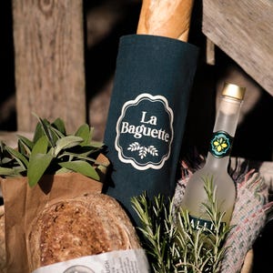 May include: A rustic picnic basket filled with a baguette in a teal holder labeled "La Baguette", a loaf of sourdough bread, a bottle of clear liquid, and sprigs of rosemary. The basket is lined with a patterned blanket.