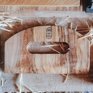 May include: A wooden handle with a cutout for a grip. The handle is made of light brown wood with dark brown grain patterns. The handle has a small, rectangular logo burned into the wood. The logo is a stylized letter "H" inside a rectangle.