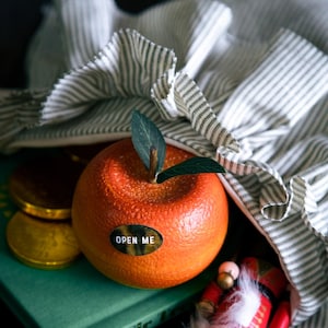 May include: A red orange with a sticker that says "OPEN ME" is sitting on a green book with gold coins and a nutcracker doll. The orange is in a white and green striped fabric bag.