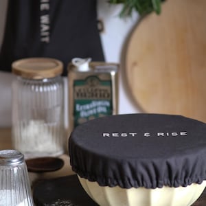 May include: A cream-coloured ceramic bowl with a black fabric lid, printed with "REST & RISE" in white. The bowl is on a wooden board, with a salt shaker and a flour jar in the background. A black apron is visible.